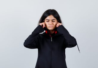 little girl plugging ears with fingers in black coat and looking bored , front view.