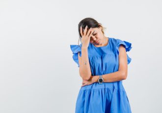 Young woman holding hand on forehead in blue dress and looking exhausted. front view.